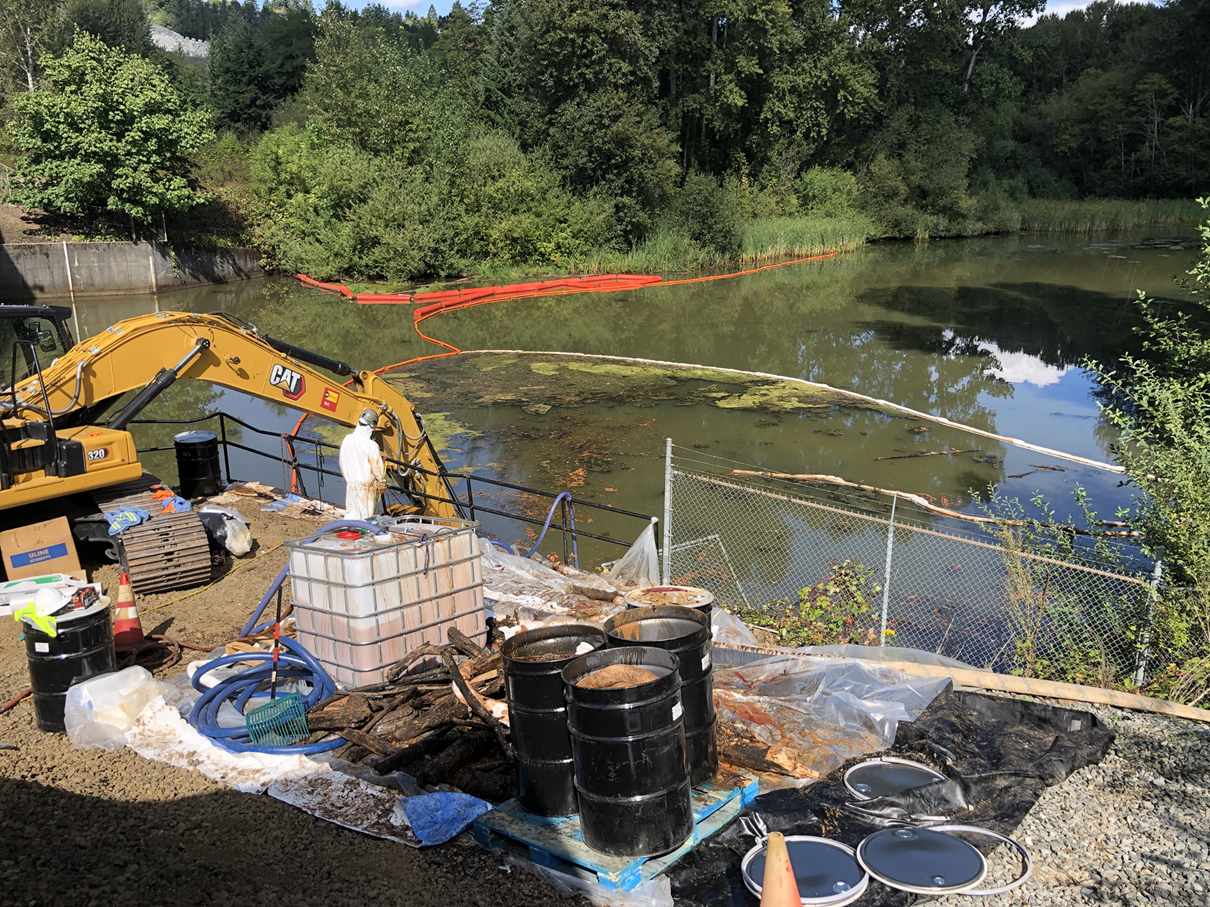 A crane bucket loader skims collected oil off a river, next to a collection of response equipment and supplies.