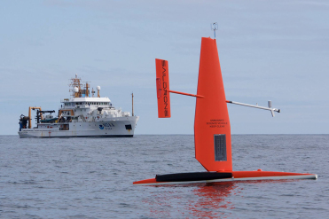A saildrone in water with a NOAA vessel in the background. 
