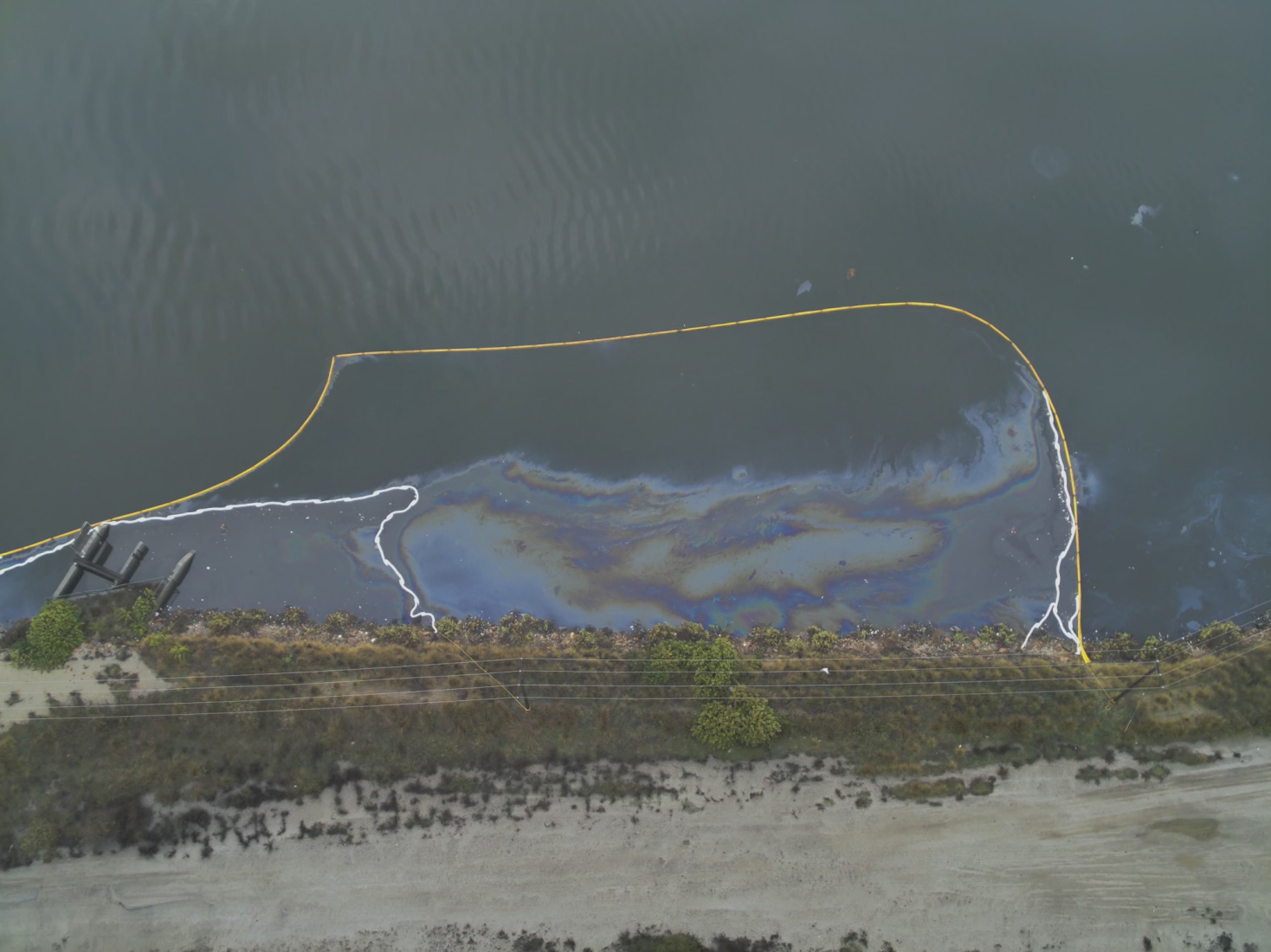 Aerial image of containment boom (yellow) and sorbent boom (white) deployed to capture oil and sheen discharging from the stormwater outfall in Dominguez Channel. Rainbow sheens are clearly visible.