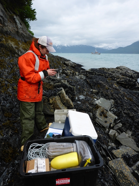 A woman with boxes of field gear on a rocky shoreline. 