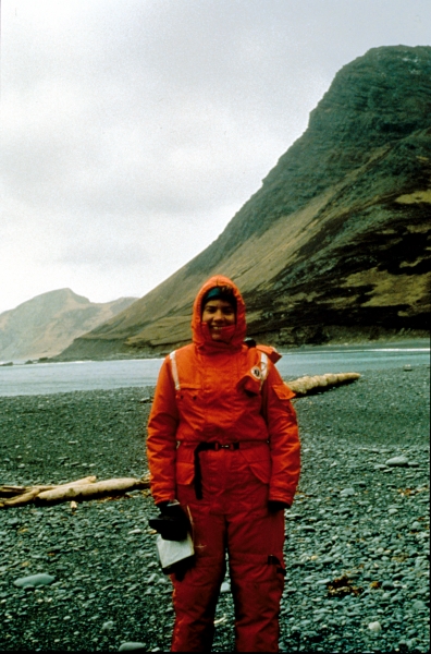 A woman in orange response gear on a rocky beach with a mountain in the background.