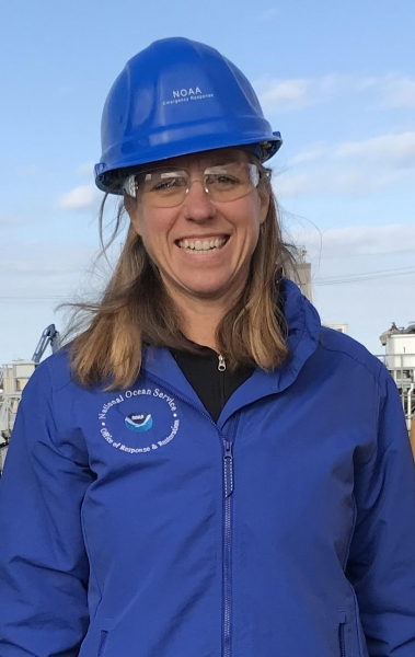 A photo of a woman wearing a hard hat and a blue jacket with the National Ocean Service logo on it. 