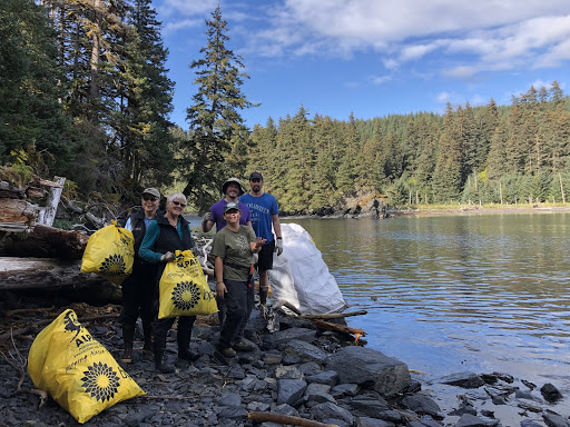 A group of people holding yellow bags on a shoreline lined with evergreen trees.