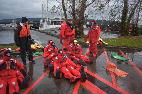 A group of people in red survival suits lined up together sitting on concrete with a dock in the background.
