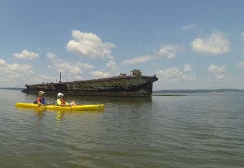 Two people in a kayak paddling in front of a wooden vessel. 