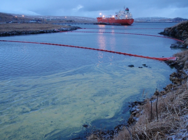A yellow substance in the water with several lines of pollution boom and a large vessel visible in the background.