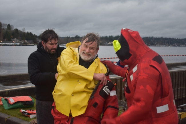 Two people help a man in a yellow rain slicker put on a survival suit