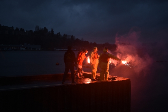 Three people holding flares at the end of a dock. 