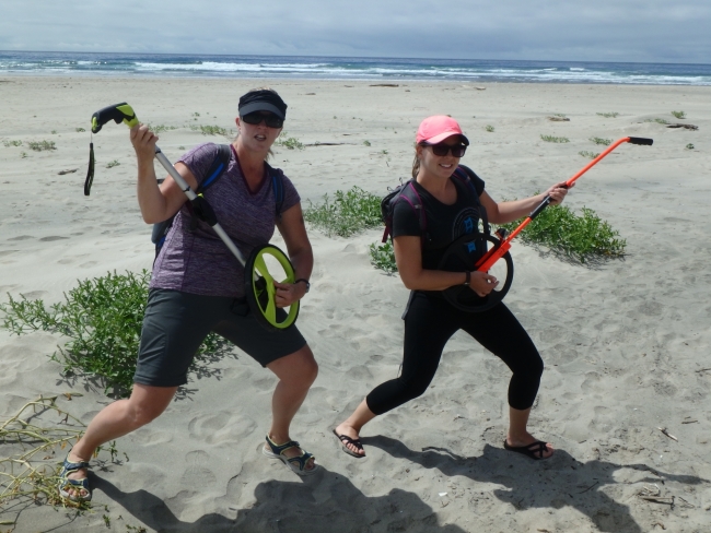 Two women strumming surveyor wheels like guitars on a beach.