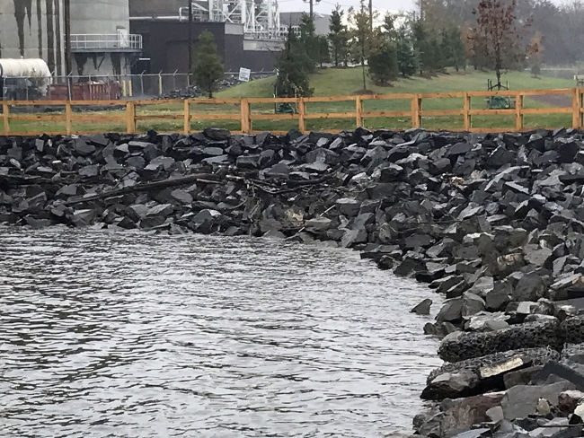 A rocky shoreline with a park and an industrial area sitting behind a wooden fence. 
