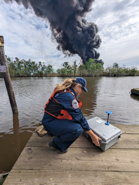 Responder sets up a box of monitoring equipment on a dock in the foreground. Large black smoke plume observed in the background of a marshland landscape.