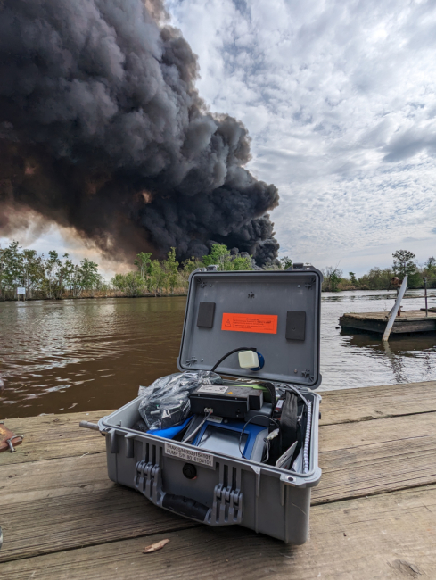 A box of monitoring equipment is positioned on a dock with a large, dark smoke plume observed in the background amongst a marshland landscape.