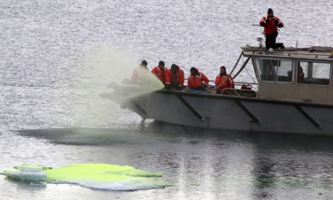 A group of people in orange gear spraying a green liquid from a boat and into the water.