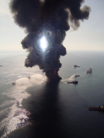 Smoke plumes rising from a structure in the water surrounded by vessels. 