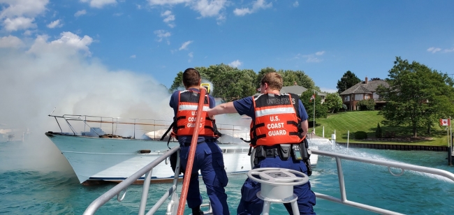 Two people in life jackets spray water at a vessel on fire.