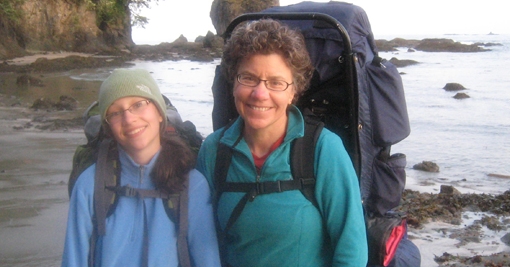 Two women with large hiking packs on posing in front of a body of water.