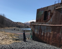A man on a beach looking at an abandoned barge. 
