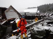 A woman in response gear with a collapsed structure covered in snow behind her.