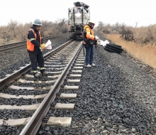 Two people in response vests and hardhats holding sorbent material and examining an area of oiled rocks next to railroad tracks.