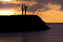 Two people standing on a rocky outcrop, fishing; backdrop is the sunset.