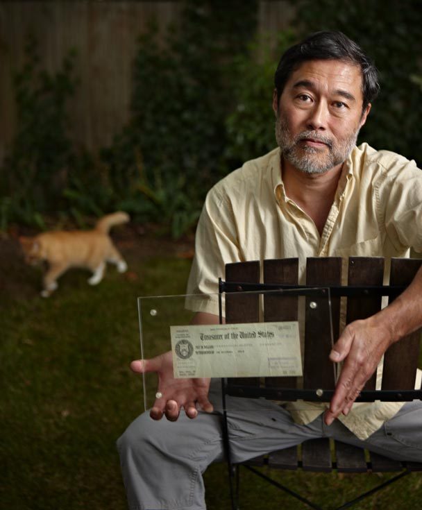 A man sitting on a chair holding a framed check with a cat in the background.