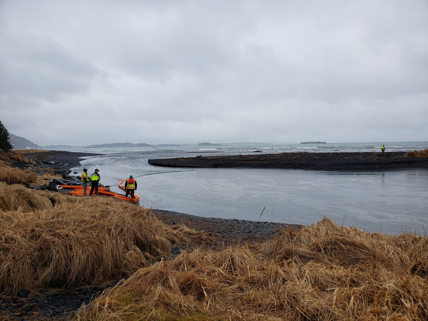 Response personnel on a shoreline near a body of water with visible oil sheen.