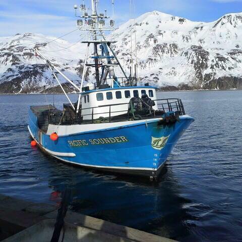 A fishing vessel with snow-covered mountains in the background.