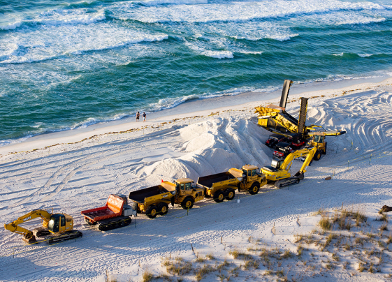 Several trucks and other heavy machinery lined up on a sanding beach with ocean waves in the background. 