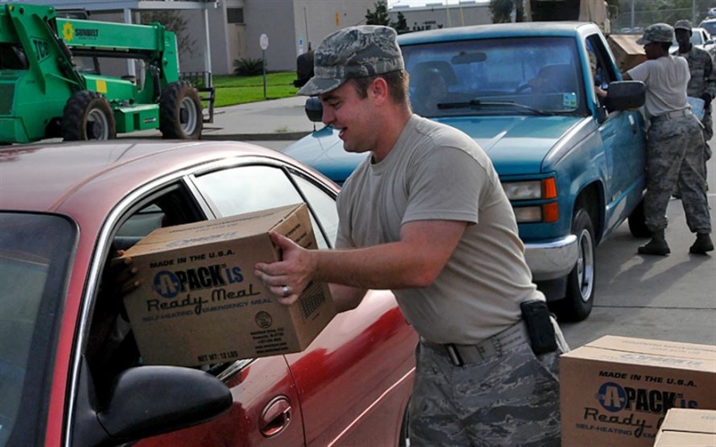 A uniformed military soldier handing a box labeled "Ready Meals" through a car window.
