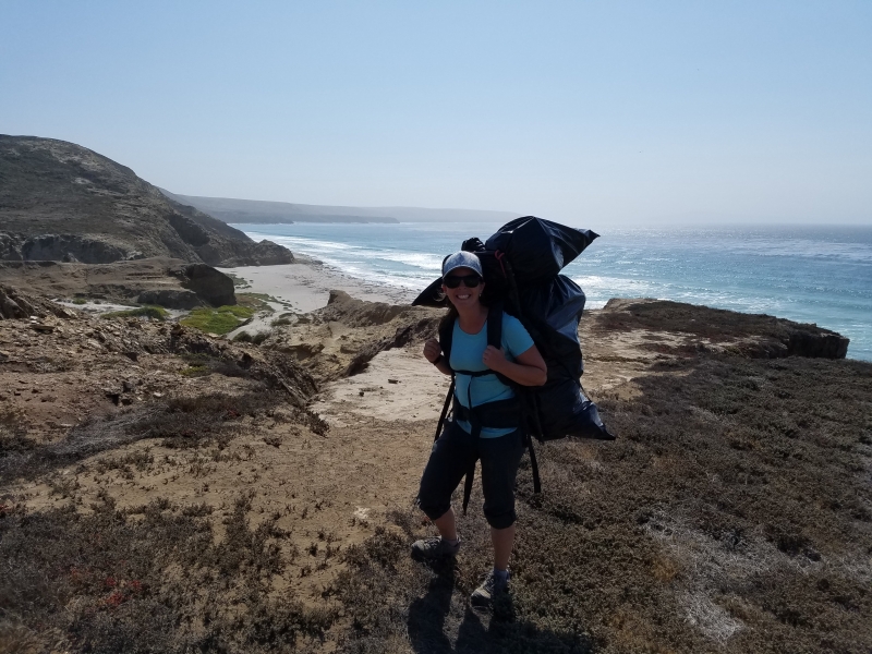 A woman with a large backpack on a hilly shoreline area. 