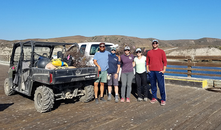 A group of people on a boardwalk posing for a photo next to a vehicle with a pile of marine debris in the truck bed.