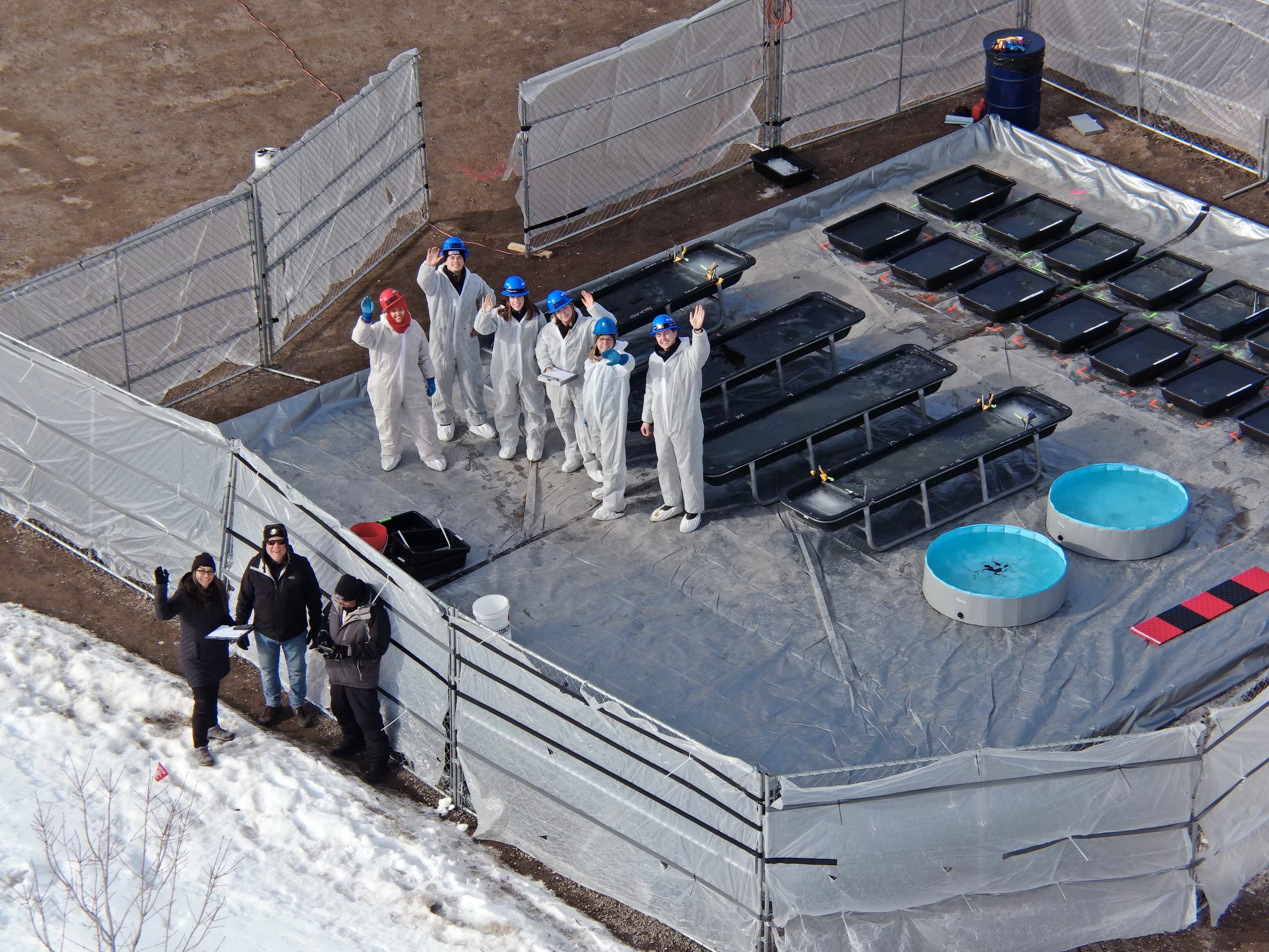 Aerial view of a group of oil spill specialists in PPE waving at the drone taking the shot. The group stands next to plastics totes staged during a field experiment in organized rows, each containing treatments of oil and diesel. The surrounding environment is icy.