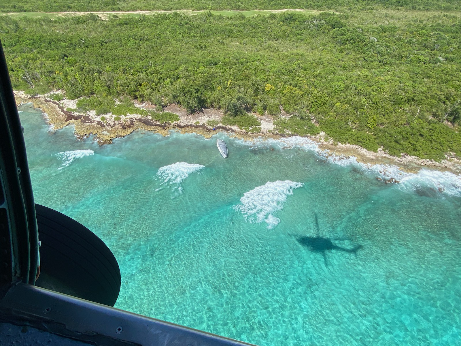 Aerial view from helicopter of a vessel grounded in blue-green water, near vegetated shoreline.