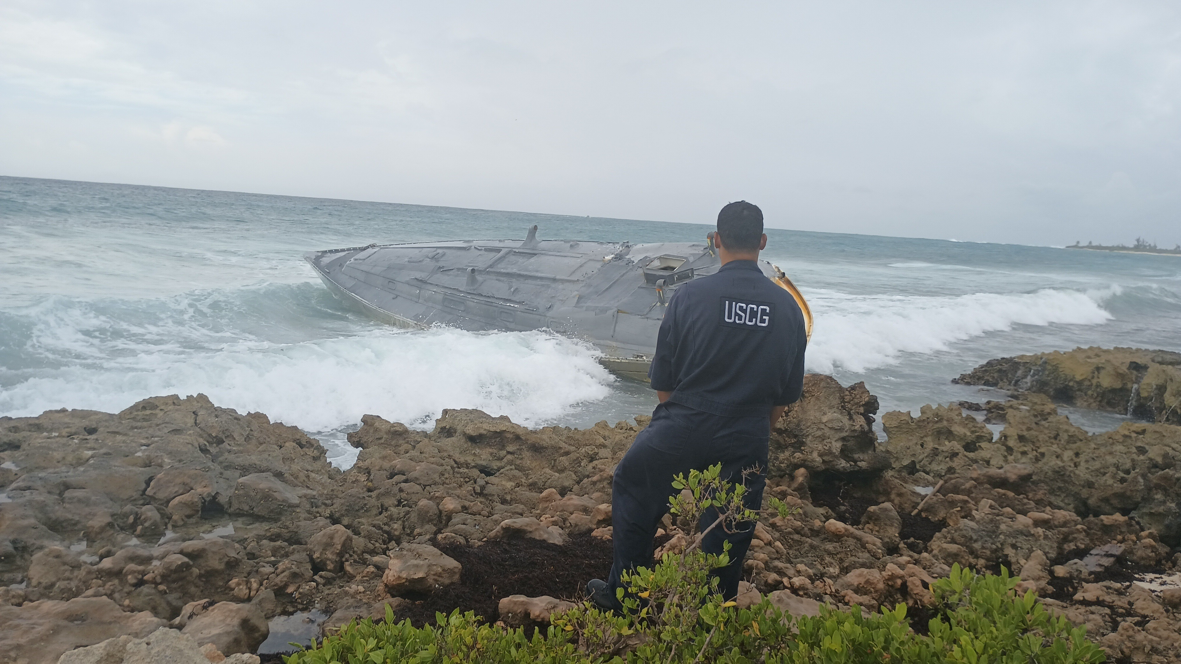A USCG spill responder stands on a rocky shoreline, looking at a semi-submersible vessel that has grounded nearby.