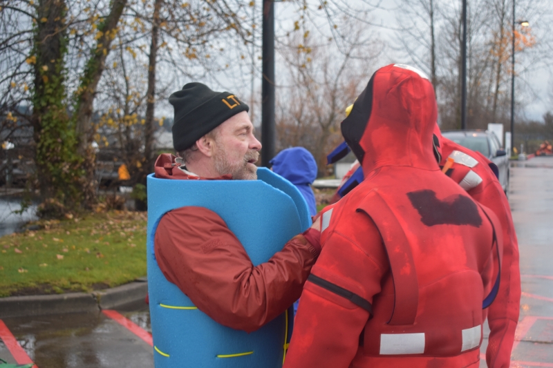 A man in in a blue life preserver speaking to two people in survival, or gumby, suits.