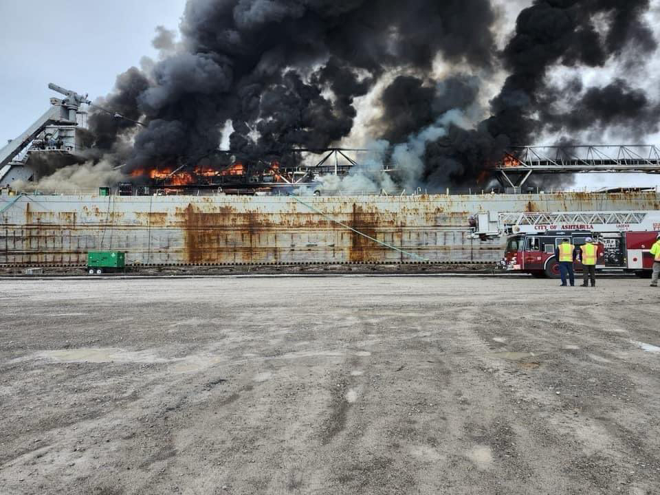 Large bulk freighter on fire while docked in a port. Firefighters stand beside a fire truck next to the vessel.