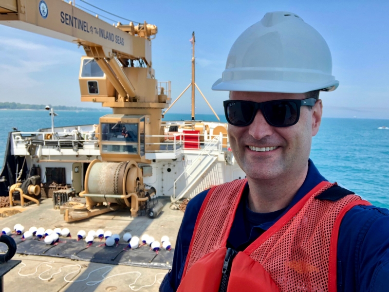 A man in a hard hat and an orange vest with large equipment and a shoreline in the background. 