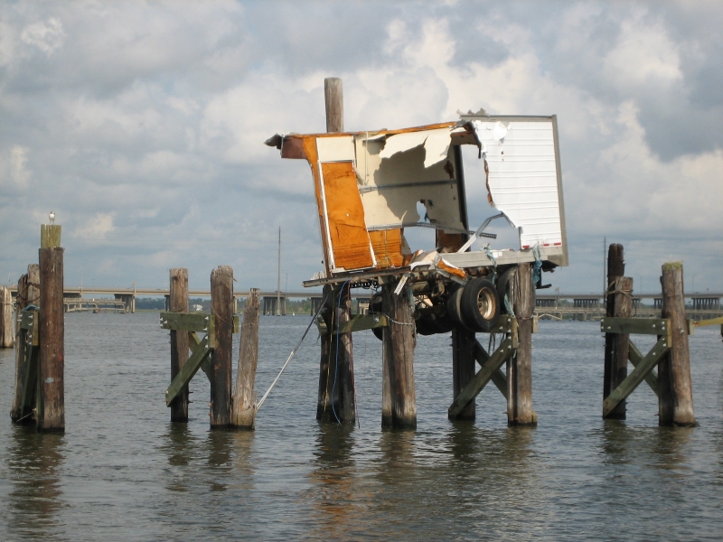 A part of a semi-truck trailer on the remains of a damaged dock above the water.