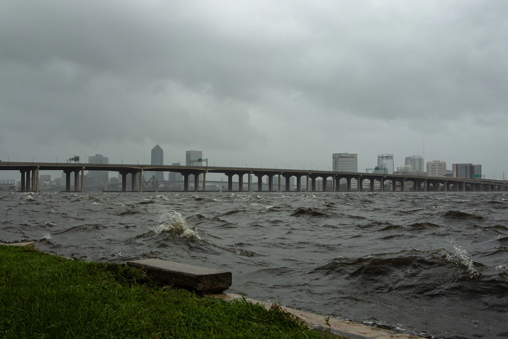 A city skyline and bridge are observed in the background, while a stormy river churns in the foreground.