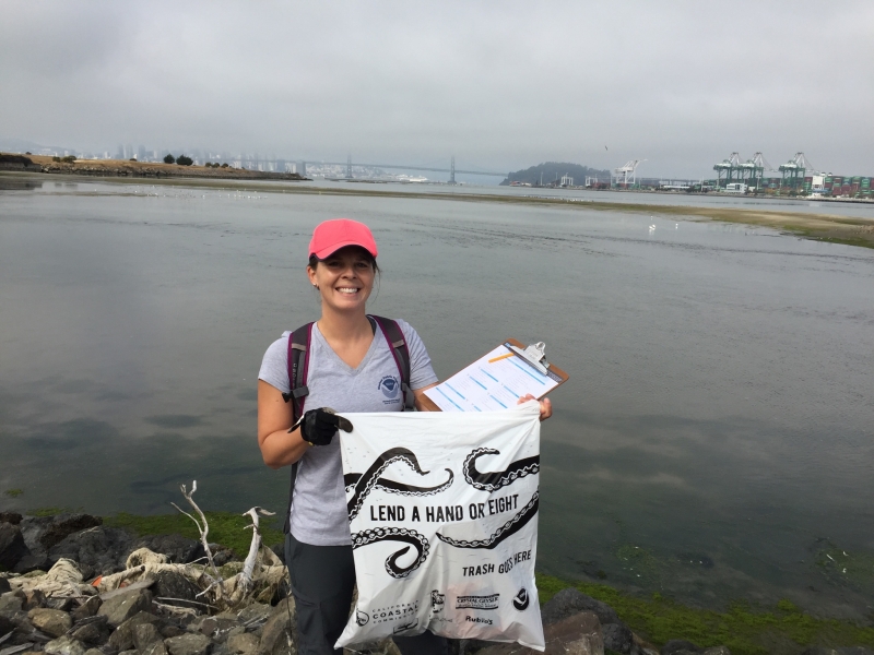 A woman holding up a plastic bag that says "lend a hand or eight" with a body of water and an industrial shoreline in the background.