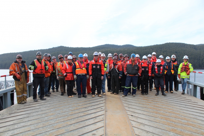 A group of people in hard hats and orange jackets standing on a dock with mountains in the background. 