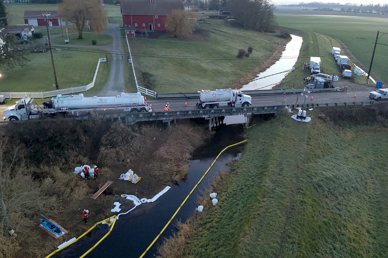  The Olympic Pipeline spill emergency response in Conway, WA. Photo shows Hill Ditch and SR 534 looking south. The boom in the lower left of the photo is capturing the spilled gasoline for collection by a skimmer connected to the vacuum trucks parked on the bridge.