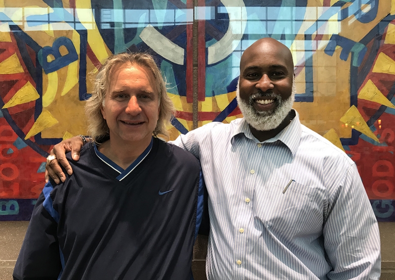 Two men standing side by side with their arms around each other as they pose in front of a painting. 