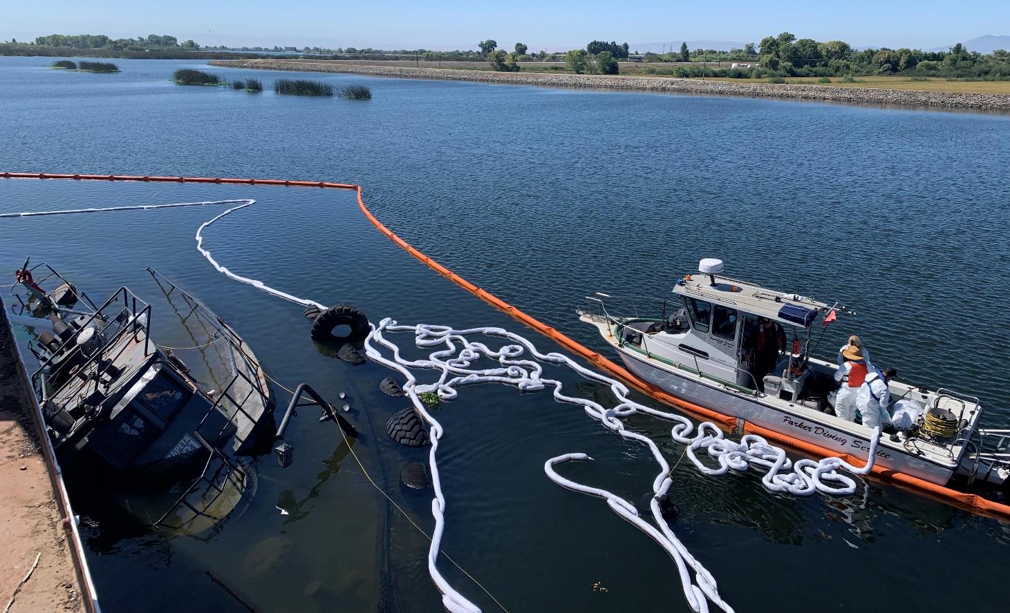 Response workers on a vessel distribute sausage boom near a sunken tug in a river.