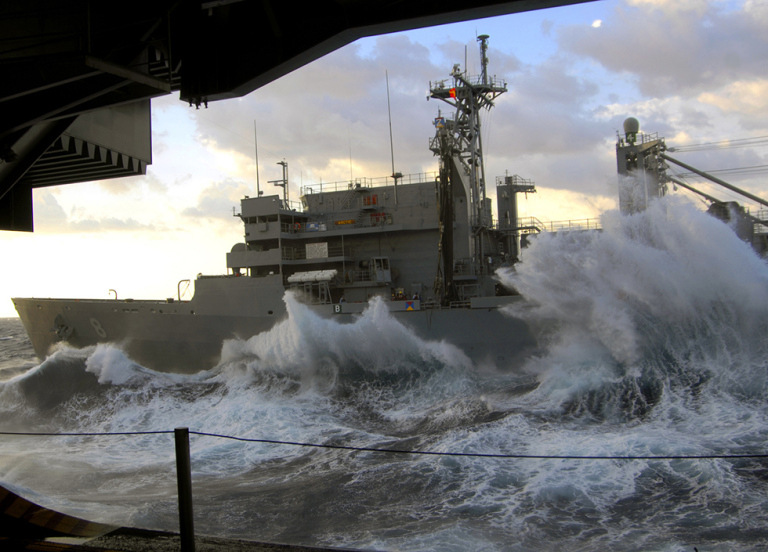 A photo of a marine vessel with waves crashing around it taken from another vessel. 