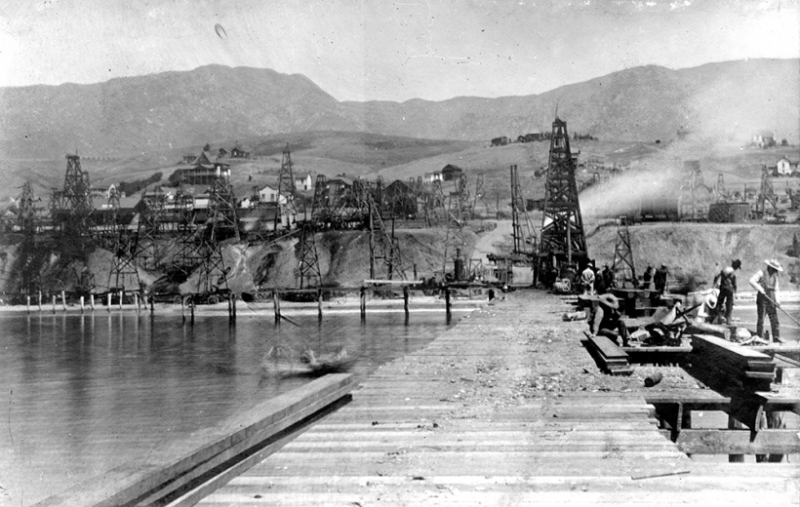 A view looking down a wharf toward shore with an oil field in the background. 