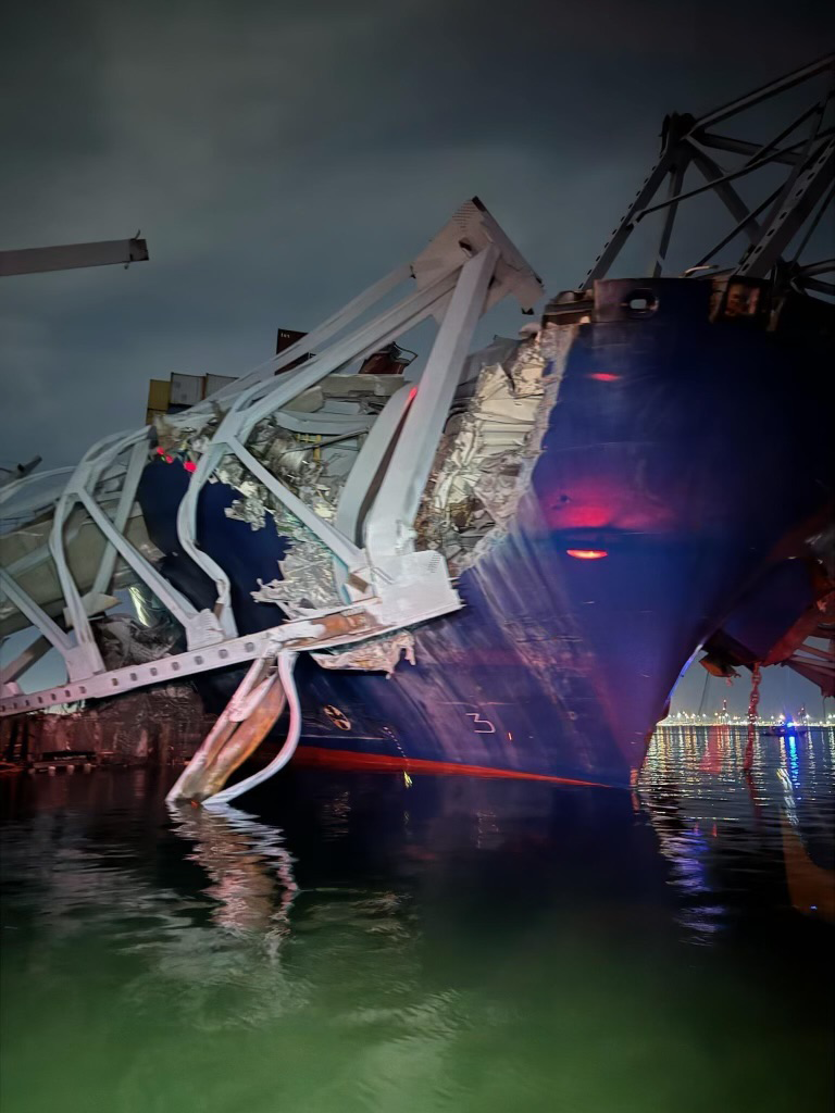 Damaged bow and containers lie underneath a section of collapsed bridge that lies across a large container ship's bow.