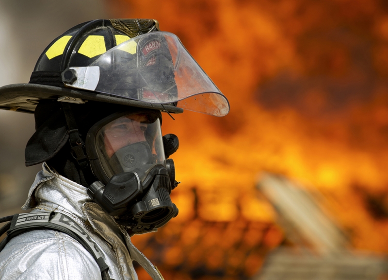 A firefighter in full gear with a fire blazing in the background. 