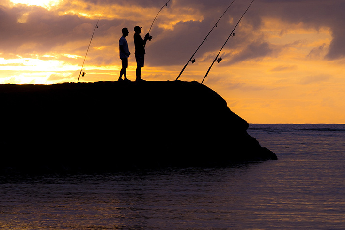 Two men fishing on a rocky outcrop with a colorful sunset in the background.