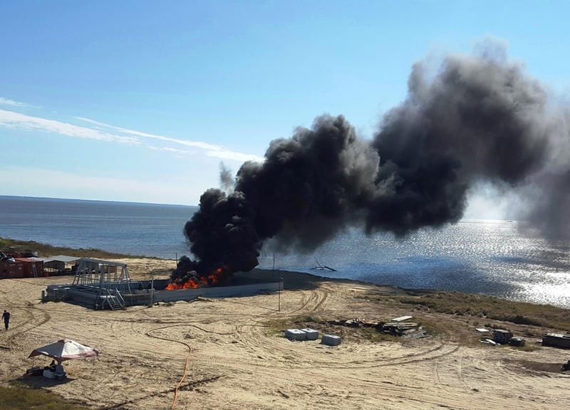 A fire with black smoke rising up from it enclosed in a structure on a sandy beach with water in the background. 